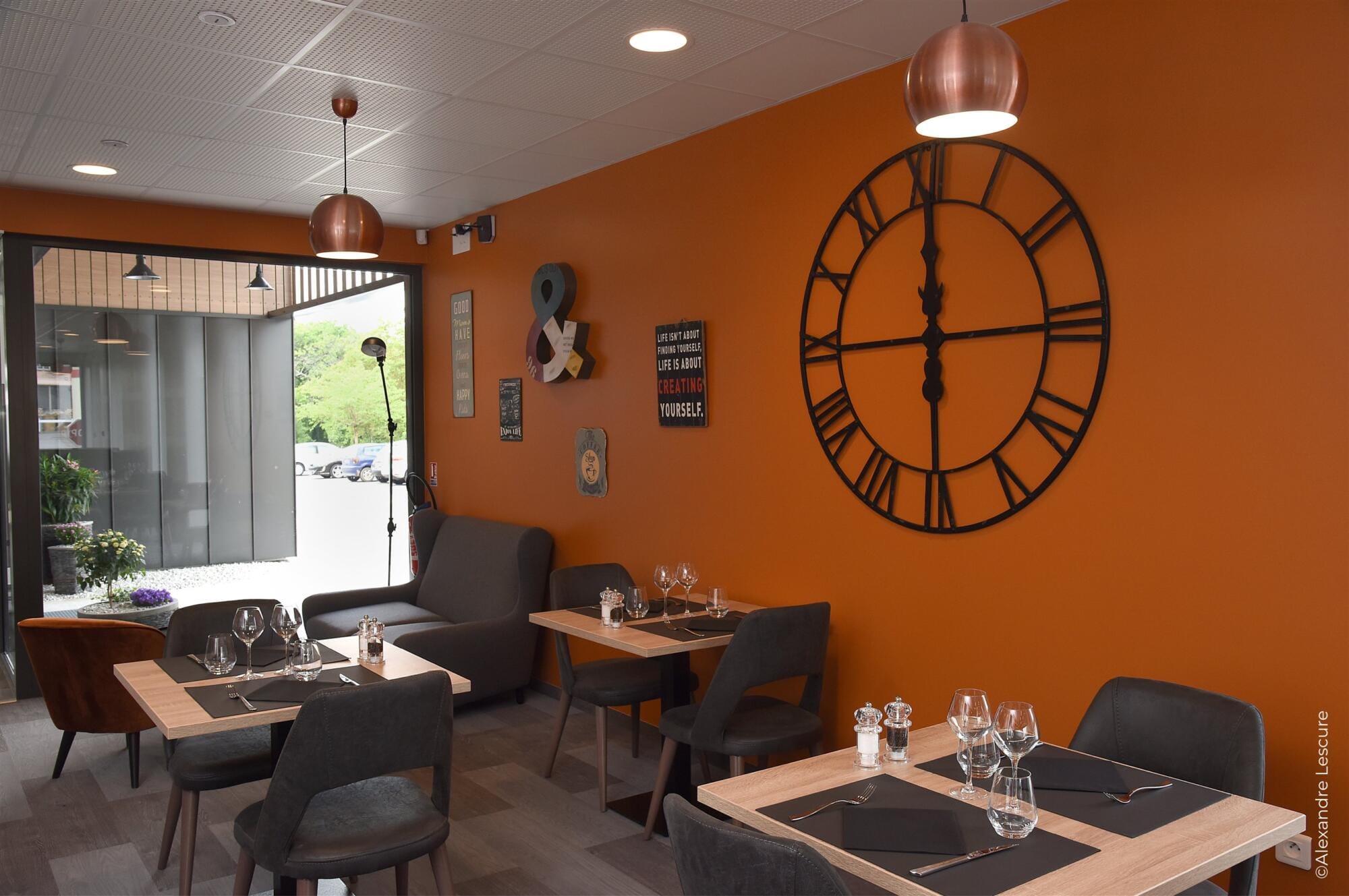 Vue salle restaurant avec tables en chêne naturel sur fond de mur orange et pendule acier noir, par Virginie Vaudenay, Décoratrice UFDI à Sancerre, dans le Cher (18), la Nièvre (58) , le Loiret (45) et L’Yonne (89), Sologne(41)